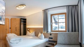 A modern hotel room with a curved bed, a seating area, a window with curtains, and a wooden wardrobe. - Vacation apartment Vergissmeinnicht - ROSENHOF Apartments & Wellness & Bauernhof (Nauders)