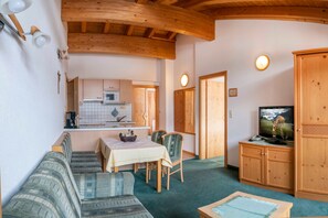 A cozy room with a wooden ceiling, a dining area with a table and chairs, a television, and a kitchen in the background.