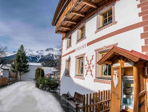 A traditional building with a wooden facade, a sign indicating a 24-hour service, and a view of snow-covered mountains in the background. - Edelweiss vacation apartment - ROSENHOF Apartments & Wellness & Farmhouse (Nauders)