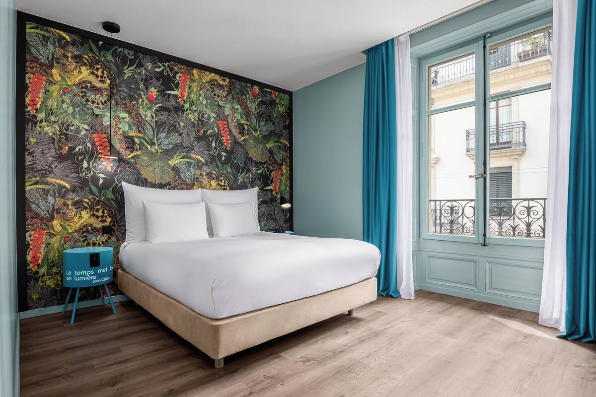 Un dormitorio con una cama grande, un mural floral en la pared, una mesita auxiliar azul pequeña y una ventana con cortinas.