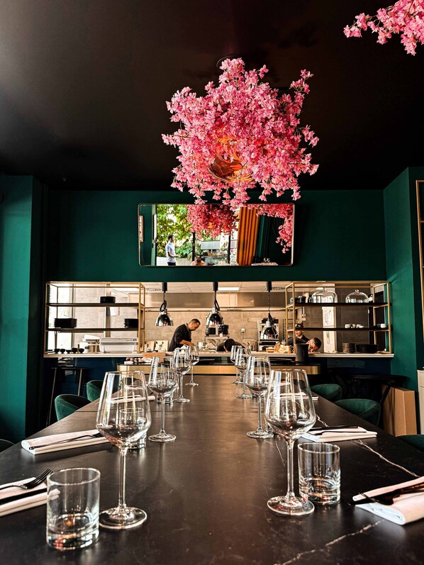 A restaurant with a long table set for dining, a chef in the background, and a large pink floral arrangement above the table. - Marmont Hotel (Geneva)