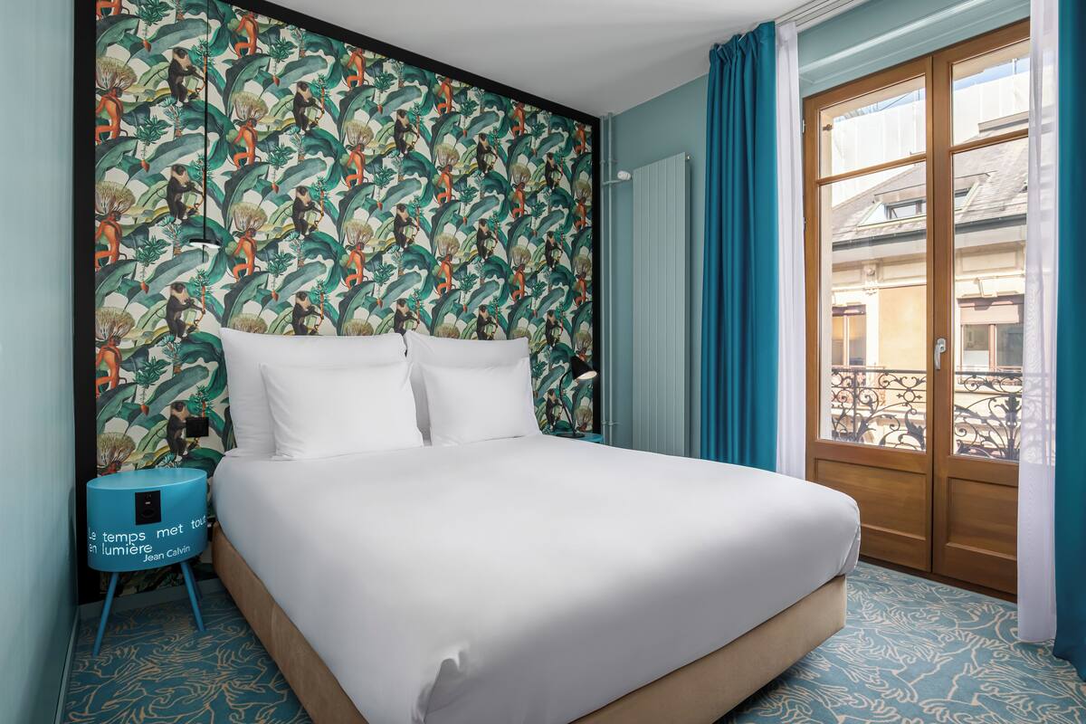 Una cama bien hecha con sábanas blancas en una habitación con diseño de papel tapiz tropical, una mesita de noche azul y vistas a un balcón a través de una puerta de madera.