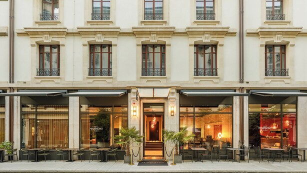 Fachada de hotel con una entrada imponente, asientos exteriores y amplias ventanas.