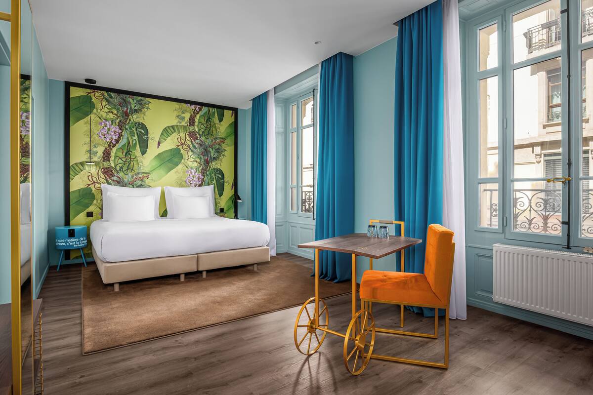 Un dormitorio con una cama grande, una mesita pequeña, una silla naranja y una ventana con cortinas azules.