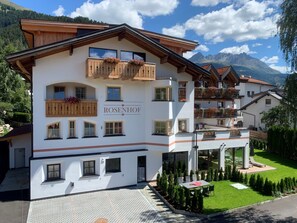 A multi-story hotel with balconies, a garden, and a mountain backdrop. - Vacation apartment Enzian - ROSENHOF Apartments & Wellness & Farmhouse (Nauders)
