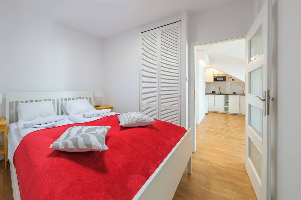 A bedroom with a bed, white walls, a wooden floor, a wardrobe, and a kitchen visible through an open door.