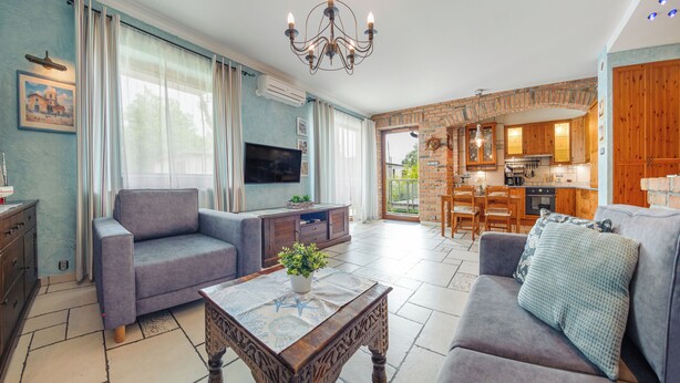 A spacious living area with a sofa, a coffee table, and a television. There is a kitchen visible in the background with wooden cabinets and a dining area. - Apartamenty Sun & Snow Willa Jazgór (Wladyslawowo)