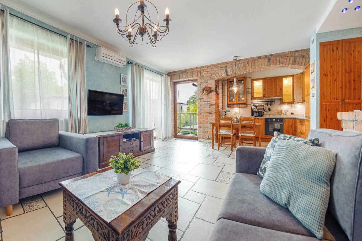 A spacious living area with a sofa, a coffee table, and a television. There is a kitchen visible in the background with wooden cabinets and a dining area.