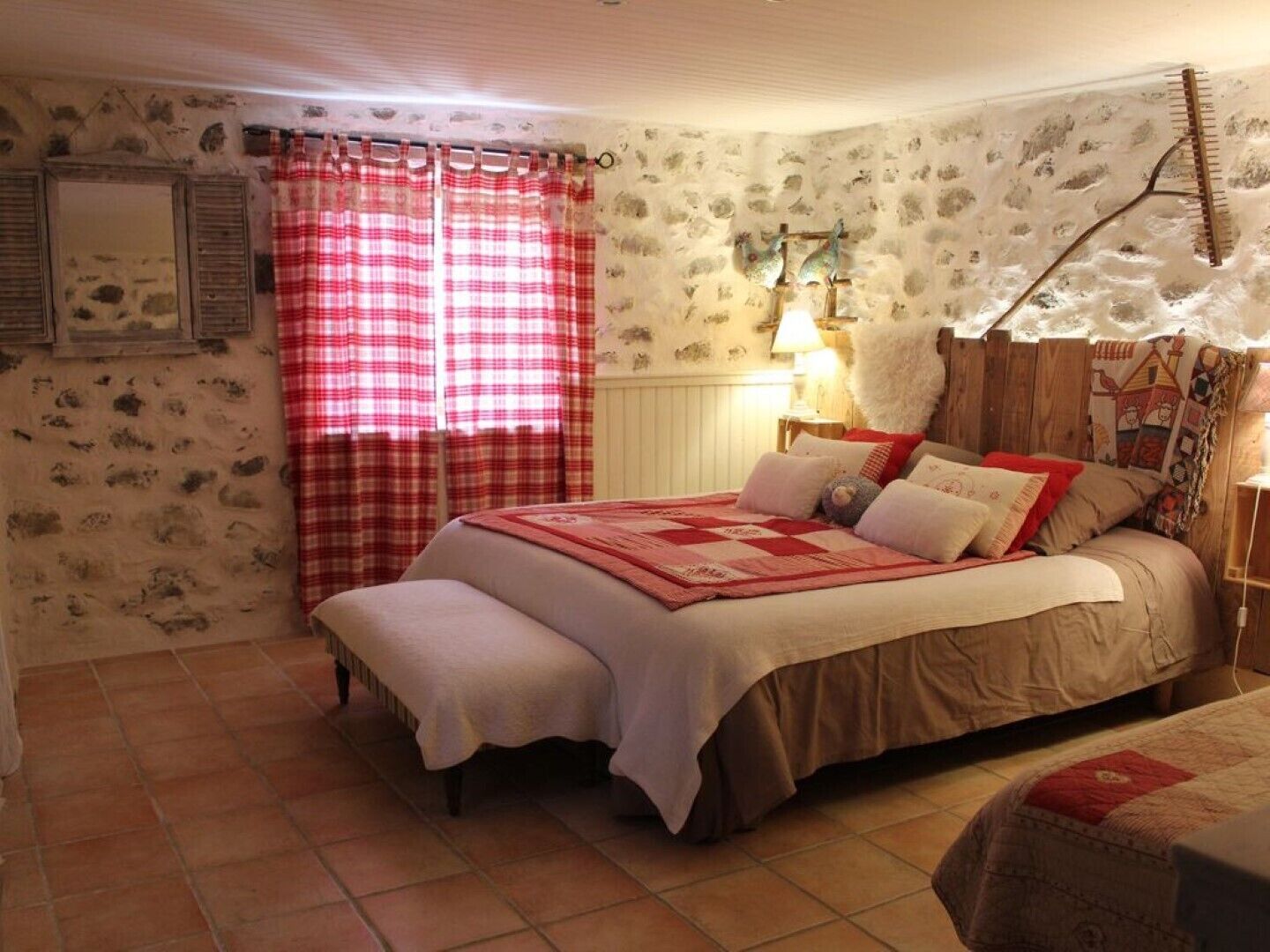 A cozy bedroom with a large bed, a bench, a wooden headboard, a red and white checkered curtain, and a wall with a rustic stone pattern.