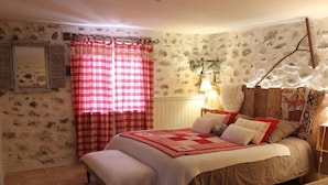 Une chambre confortable avec un grand lit, un banc, une tête de lit en bois, un rideau à carreaux rouges et blancs, et un mur orné d’un motif de pierres apparentes.