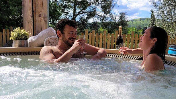 Un couple dans un jacuzzi, une bouteille de champagne à portée de main, entouré de clôtures en bois et de verdure.