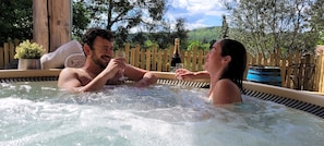 A couple in a hot tub with a bottle of champagne, surrounded by wooden fences and greenery.
