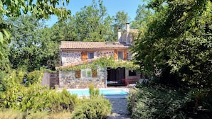 Une maison en pierre avec un toit en tuiles, une piscine et un chemin en bois menant à l’entrée.
