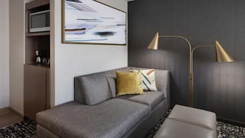 A modern hotel room with a grey sofa, a small table, a floor lamp, and a painting on the wall.