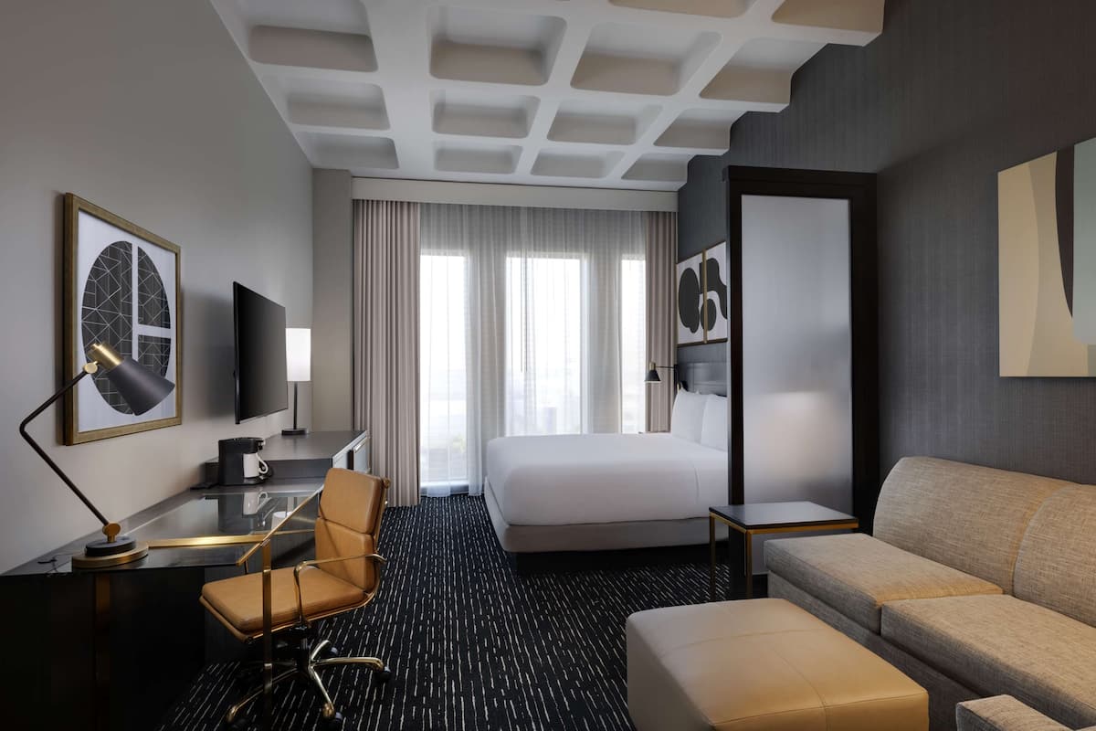 A hotel room with a large bed, a sofa, a desk with a chair, a flat-screen TV, and artwork on the walls.