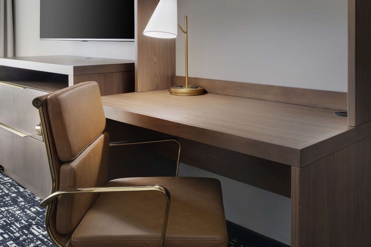 A hotel room with a wooden desk, a brown leather chair, a gold lamp, and a flat-screen TV.