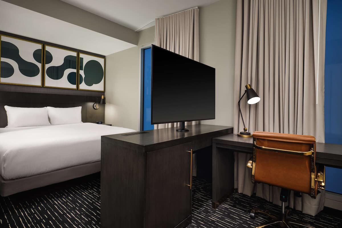 A hotel room with a bed, a desk with a TV, a chair, and a lamp.