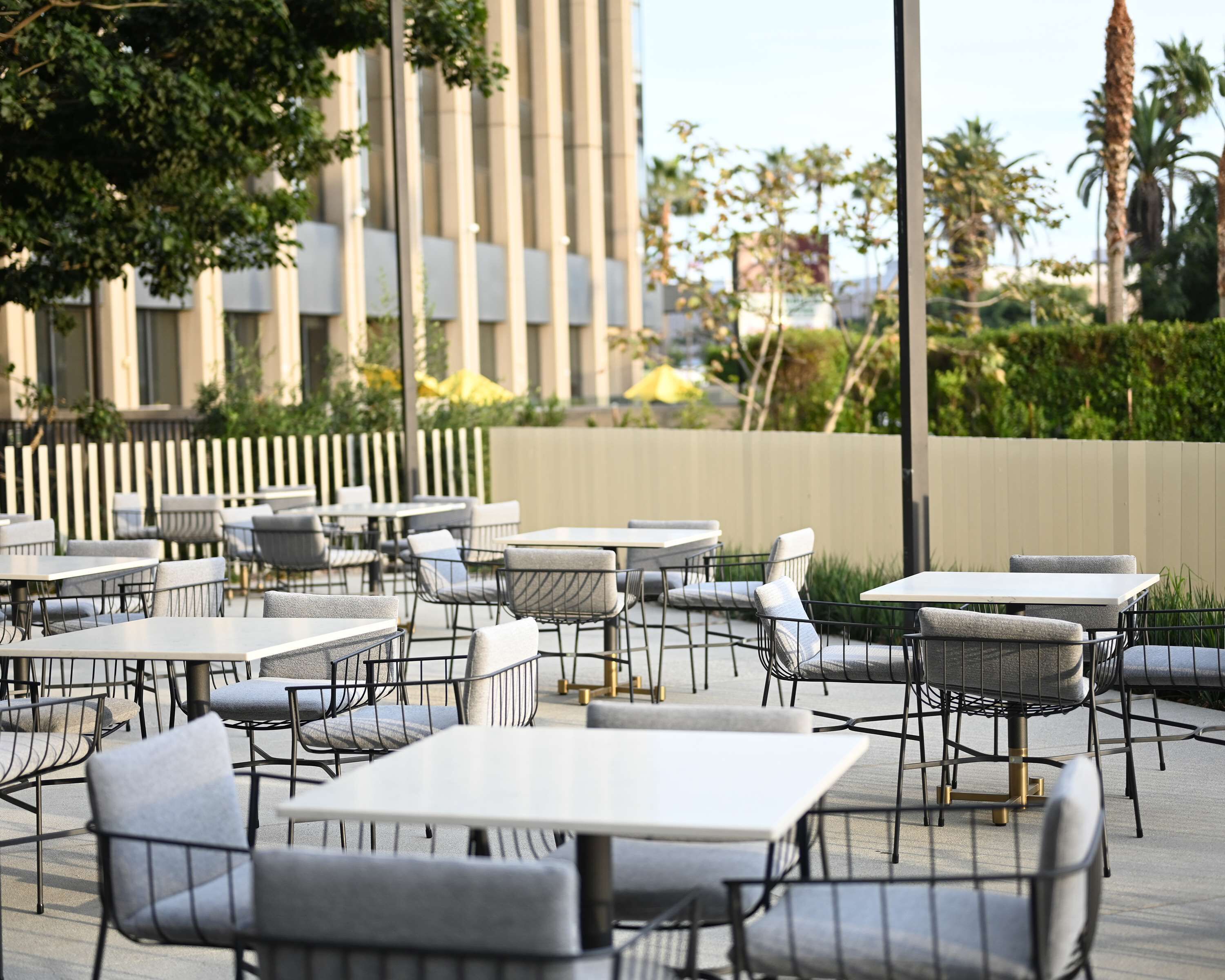 An outdoor cafe area with tables and chairs, a fence, and a building in the background.