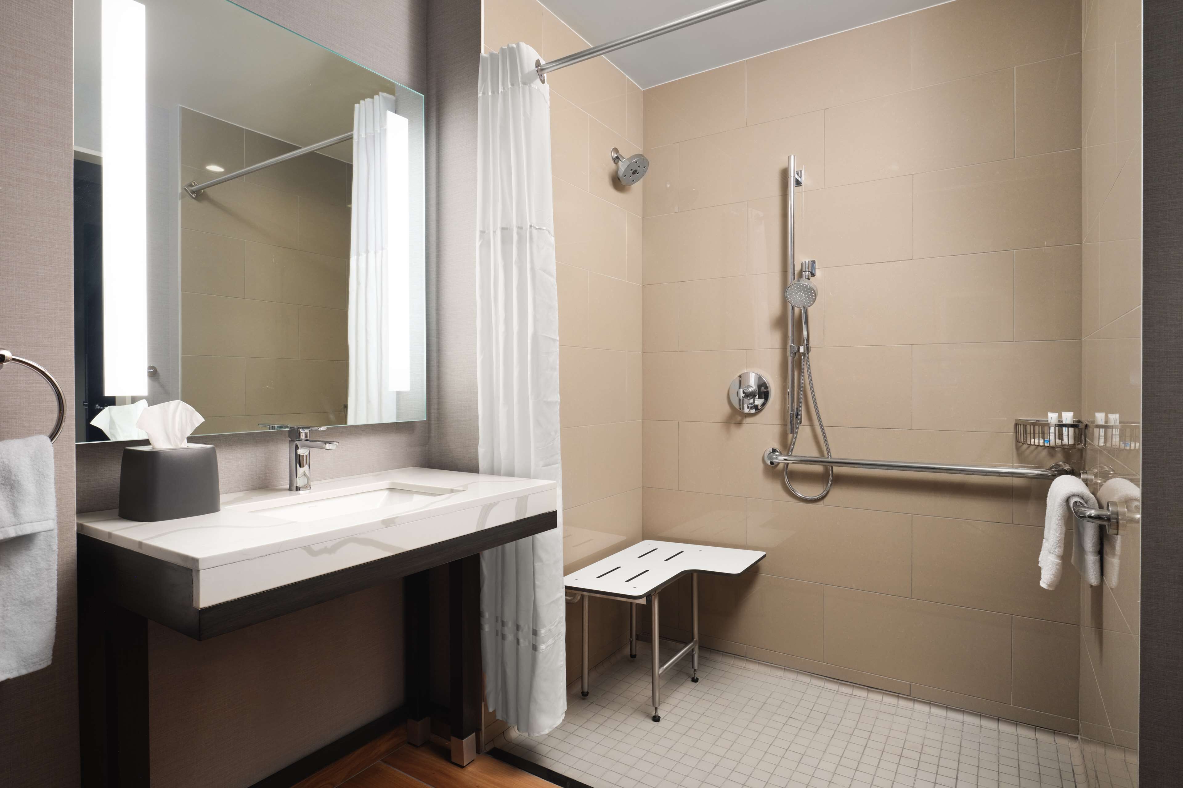 Un baño moderno con un espejo grande, un lavabo, una ducha con cabezal de ducha manual y un taburete pequeño.