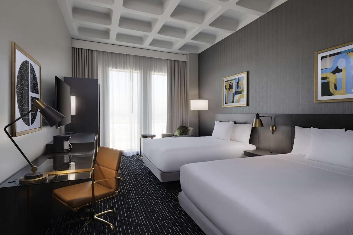 A hotel room with two beds, a desk with a chair, a TV, and artwork on the walls.