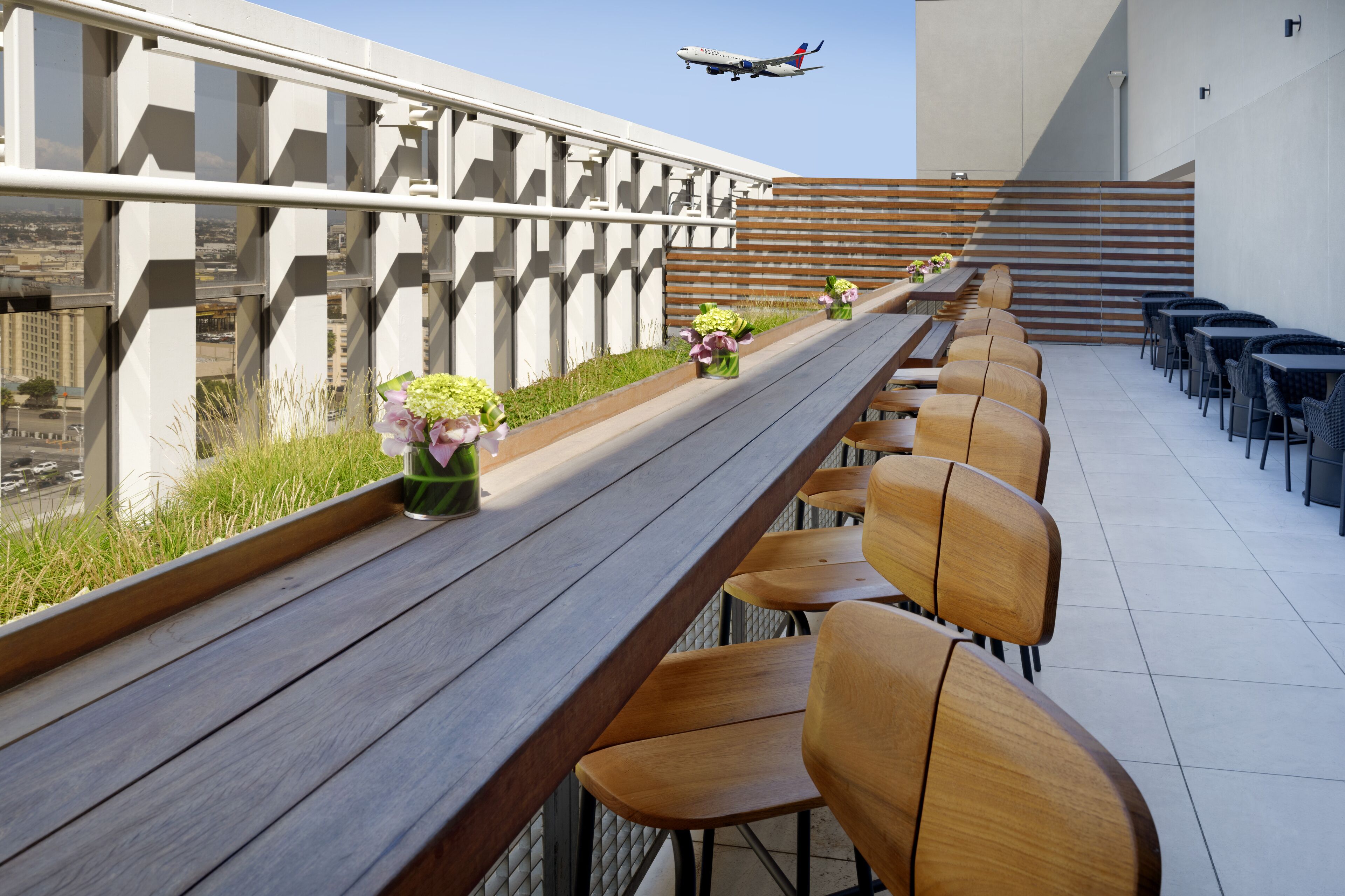 A rooftop terrace with wooden benches and chairs, a railing, and a view of a cityscape and airplane in the distance.