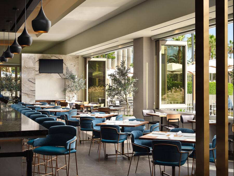 Un restaurante moderno con una barra larga, sillas azules y grandes ventanales que ofrecen vistas al exterior.