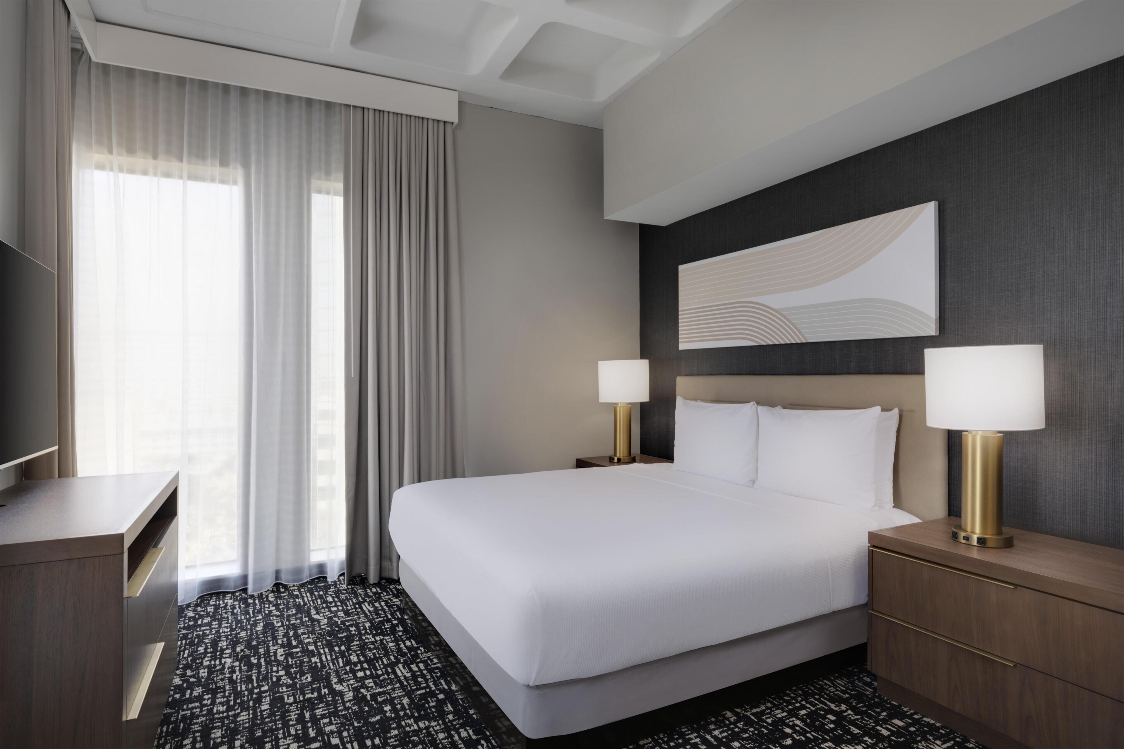 Een hotelkamer met een groot bed, twee bedlampen, een flatscreen televisie, een bureau en een groot raam met gordijnen.