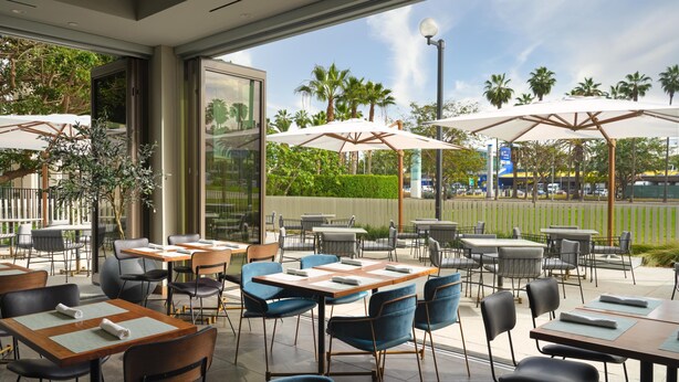 An outdoor dining area with tables and chairs, umbrellas, and a view of palm trees and a building. - Hyatt House LAX/Century Blvd (Los Angeles)