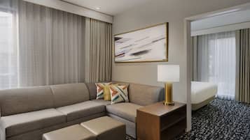 A modern hotel room with a sectional sofa, a small wooden side table with a lamp, a bed, and a large abstract painting on the wall.