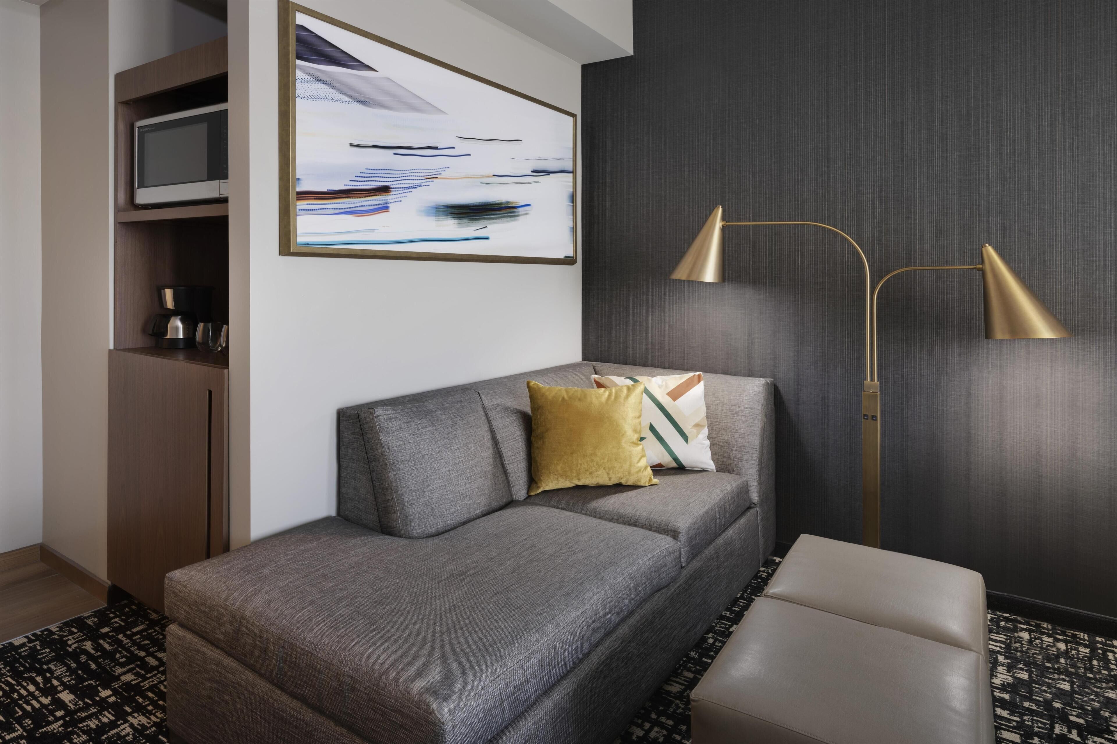 Une chambre d’hôtel moderne avec un canapé gris, un lampadaire doré, un tableau au mur et un tapis à motifs.