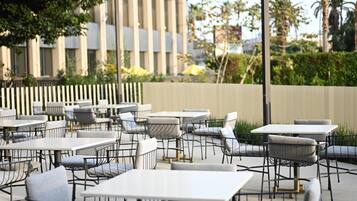 Mobiliário de café ao ar livre com mesas e cadeiras, cerca de madeira e um edifício ao fundo.
