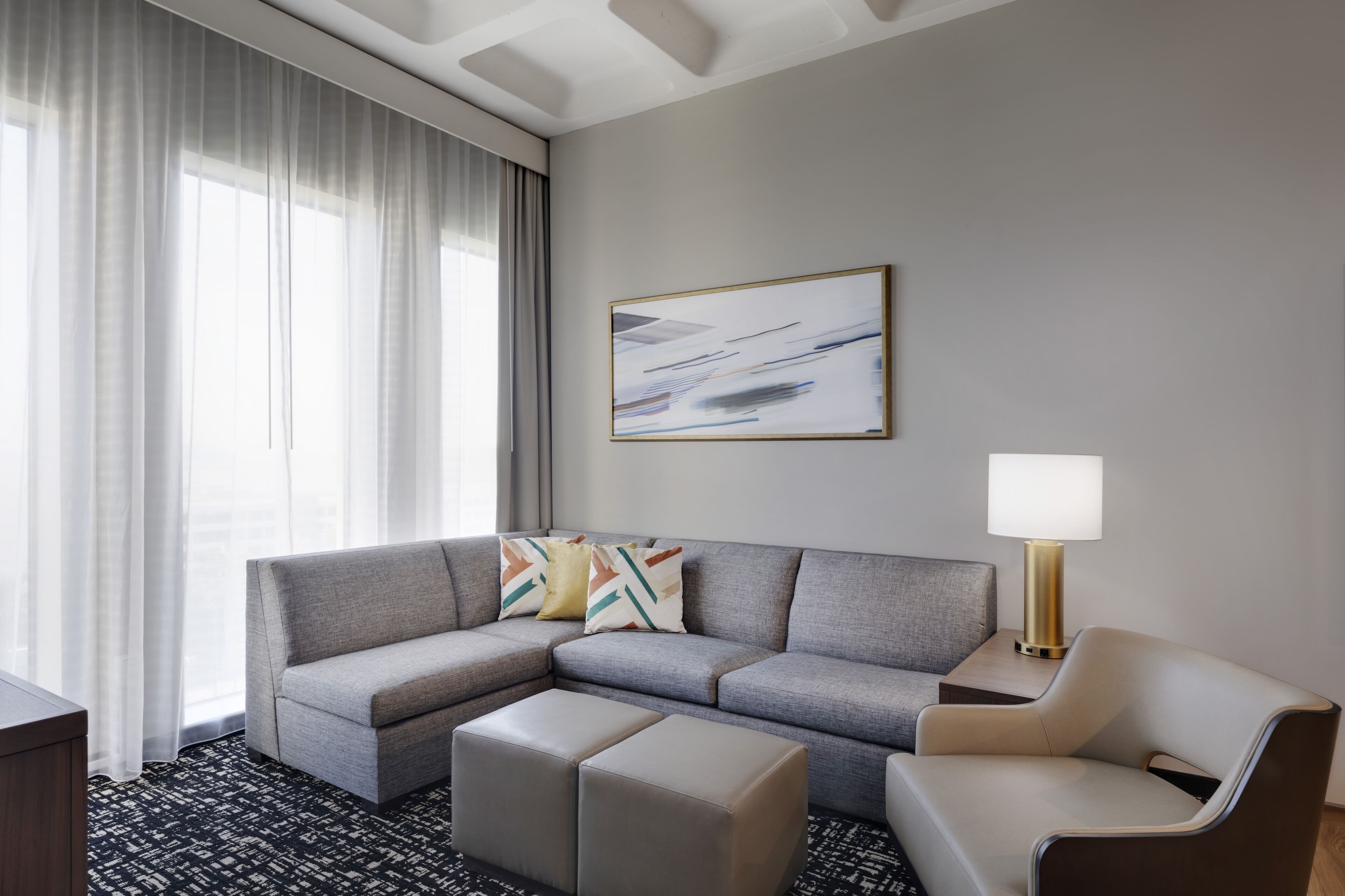 Una habitación de hotel moderna con un sofá modular gris, un sillón pequeño y un reposapiés a juego. En la pared hay una lámpara de pie y un cuadro abstracto enmarcado.