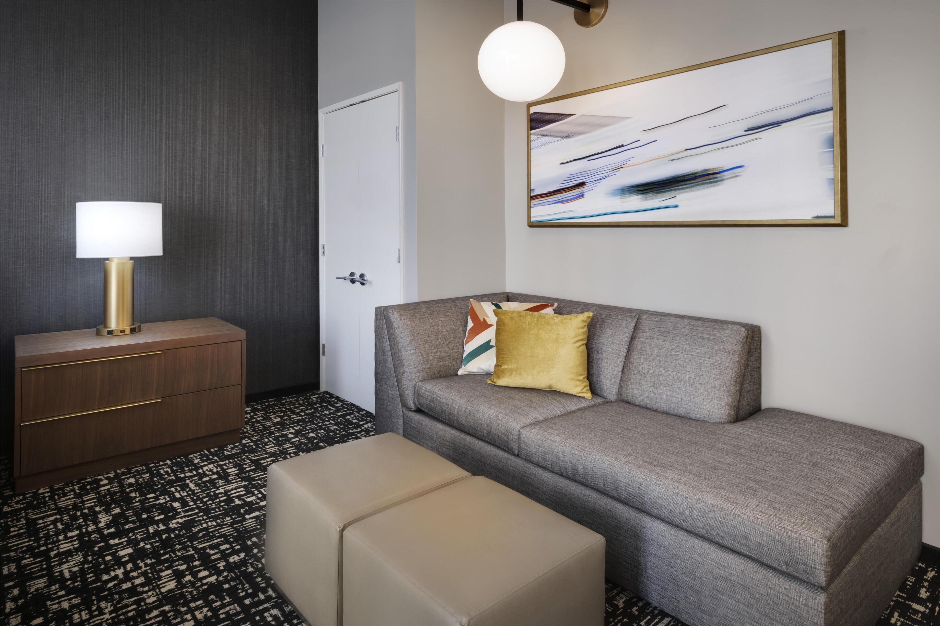 Une chambre d’hôtel équipée d’un canapé, d’un pouf, d’une table de chevet, d’une lampe et d’un tableau abstrait.