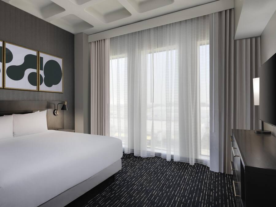 Habitación de hotel con una cama grande, un televisor de pantalla plana, un escritorio y una ventana con cortinas translúcidas.