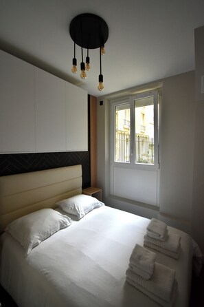 A hotel room with a bed, white linens, a headboard, a window with a view, and a ceiling light fixture.