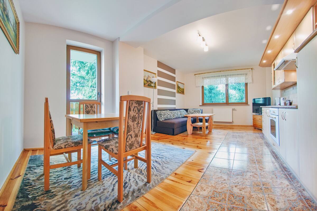 A modern kitchen with wooden cabinets, a dining area with a table and chairs, and a living area with a sofa and a TV.