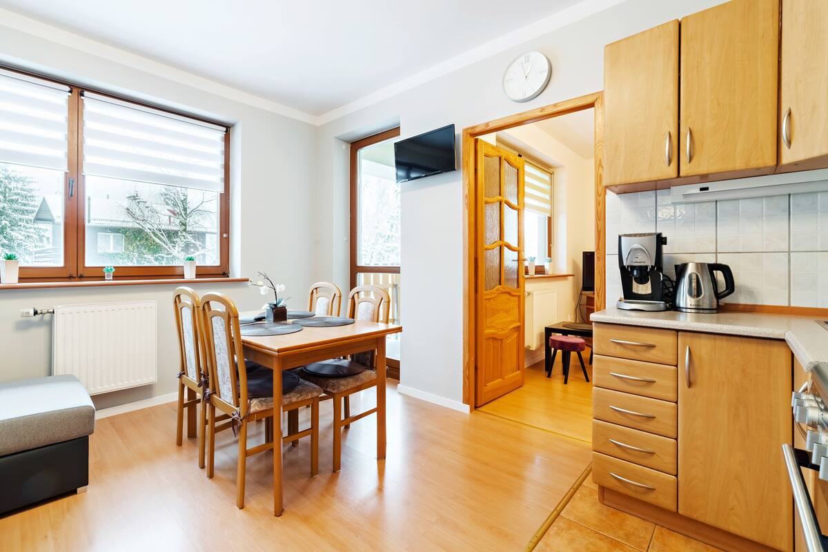 A modern kitchen with wooden cabinets, a dining table with chairs, a coffee machine, and a view of the outside through large windows.