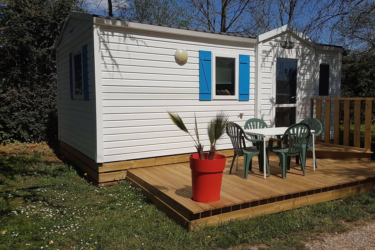 Een kleine, gelijkvloerse stacaravan met een terras, een tafel met stoelen en een bloempot.