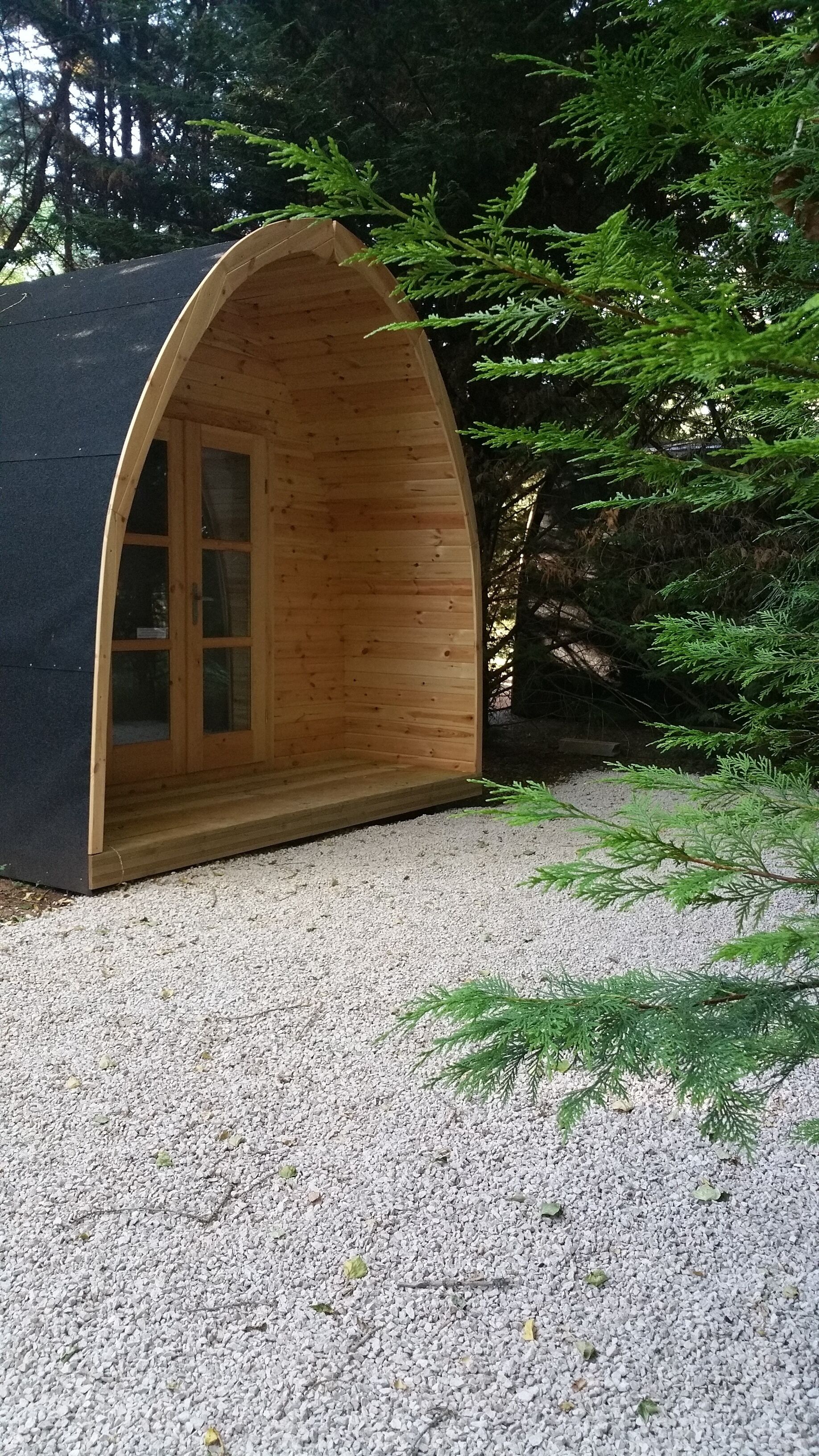 A wooden cabin with a curved roof and a single door, surrounded by gravel and trees.