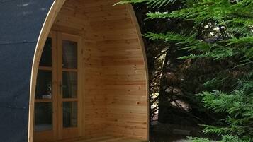 A wooden cabin with a curved roof and a single door, surrounded by gravel and trees.