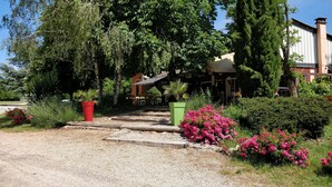 A gravel pathway leading to a building with a covered entrance, surrounded by lush greenery and flowering plants. - Au Tour De L'Aveyron (L'Hospitalet-du-Larzac)