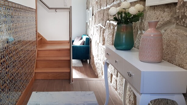 Una escalera conduce a una habitación con pared de piedra, un jarrón con flores blancas y un sofá azul.