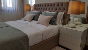 Una cama bien hecha con cabecera capitonada, dos mesitas de noche con lámparas y un televisor de pared.