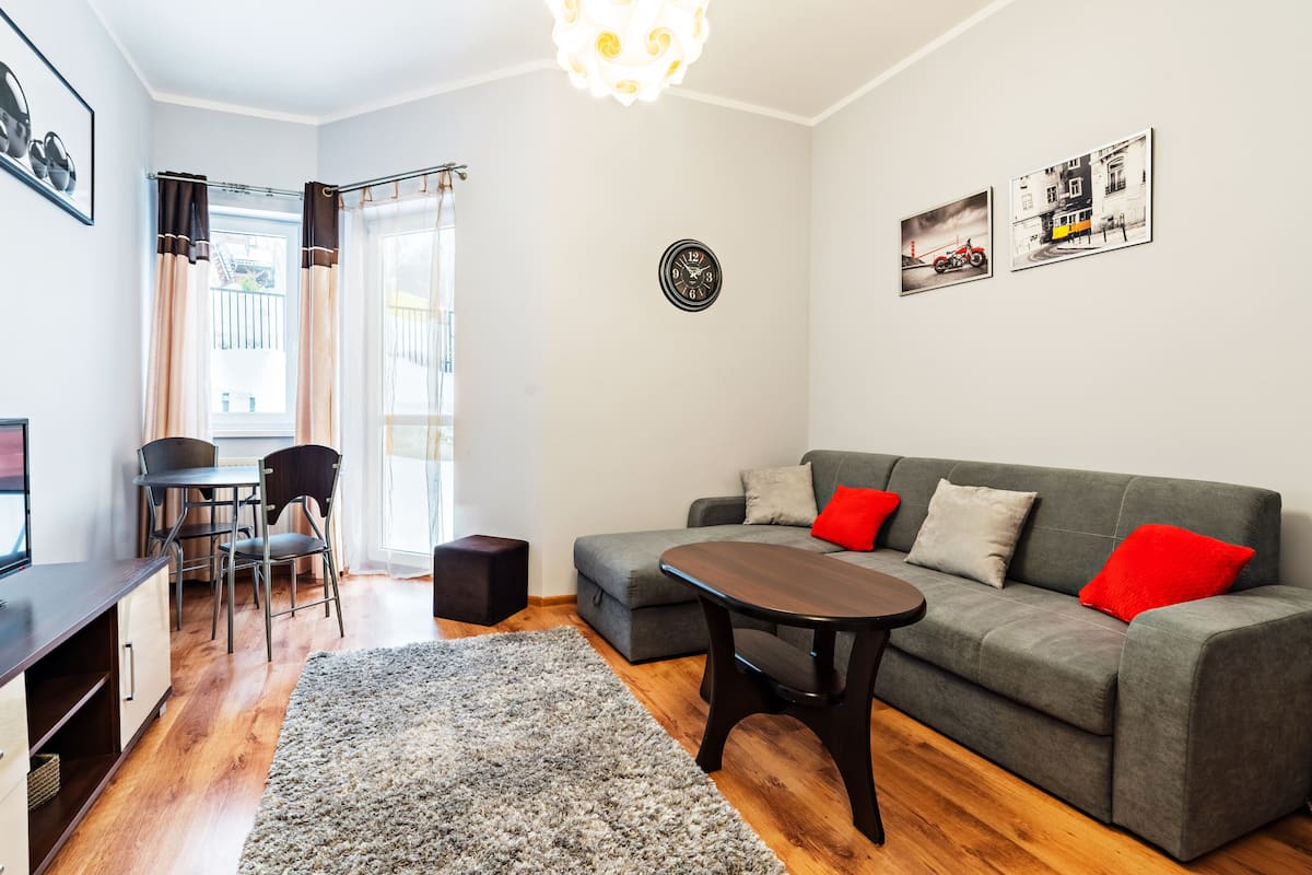 Een moderne woonkamer met een grijze bank, rode kussens, een houten salontafel, een televisie en een kleine eethoek met stoelen en een tafel.