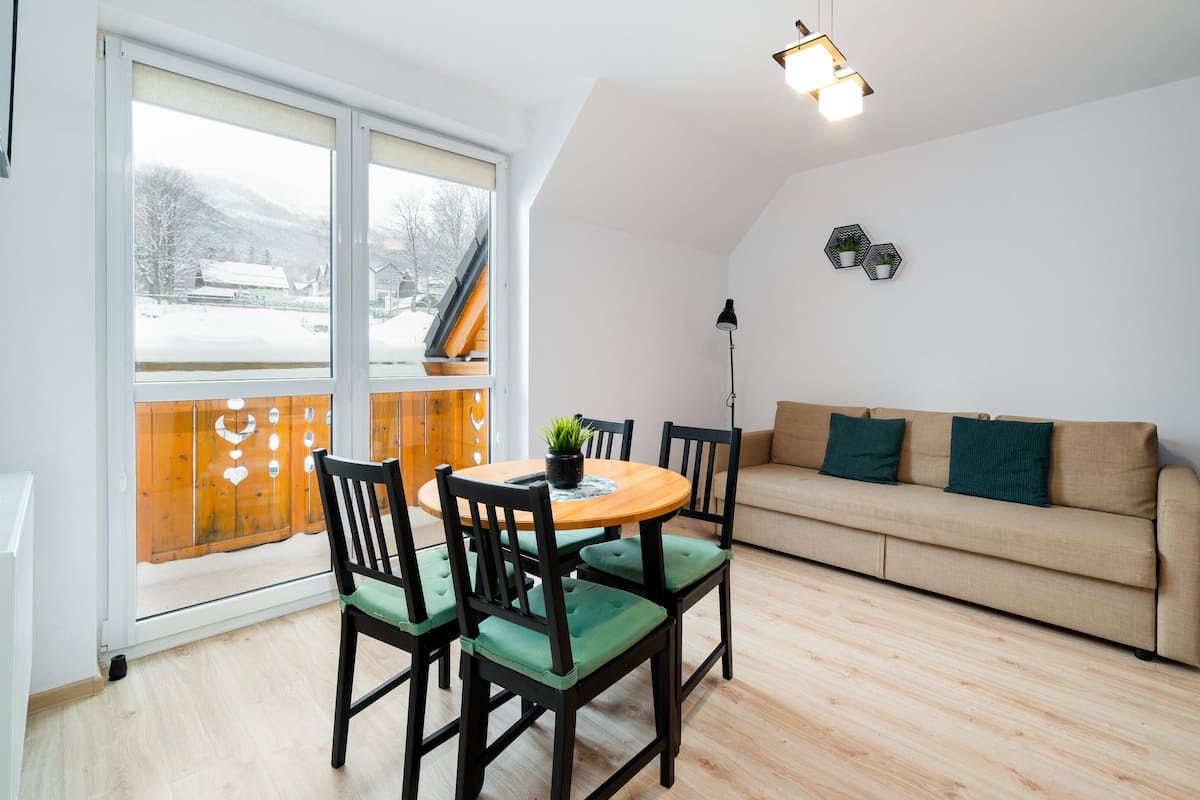 Een gezellige woonkamer met houten vloer, een beige bank en een eettafel met stoelen.