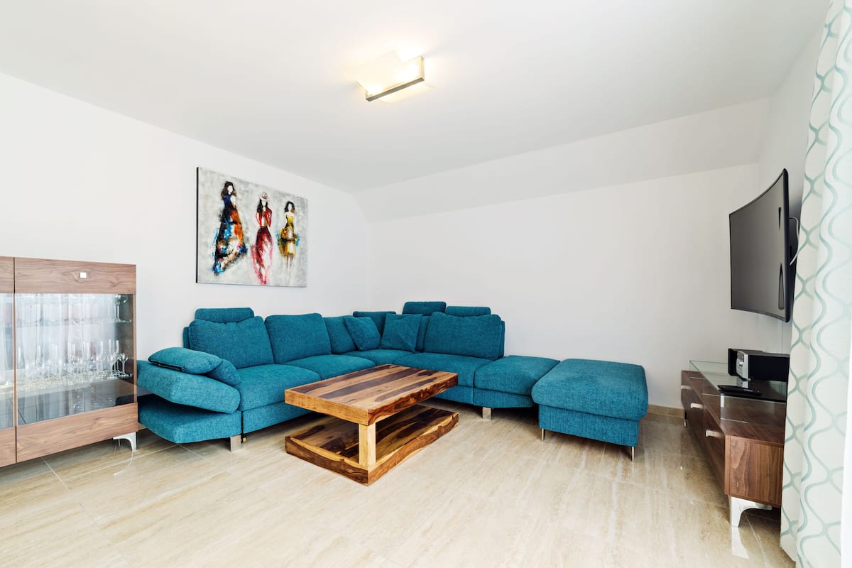 Een moderne woonkamer met een smaragdgroene hoekbank, een houten salontafel en een glazen kast met wijnglazen.