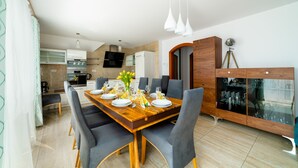 Een moderne eetruimte met een houten tafel voor zes personen, omringd door grijze gestoffeerde stoelen. Achtergrond toont een keuken met een ingebouwde oven en magnetron.