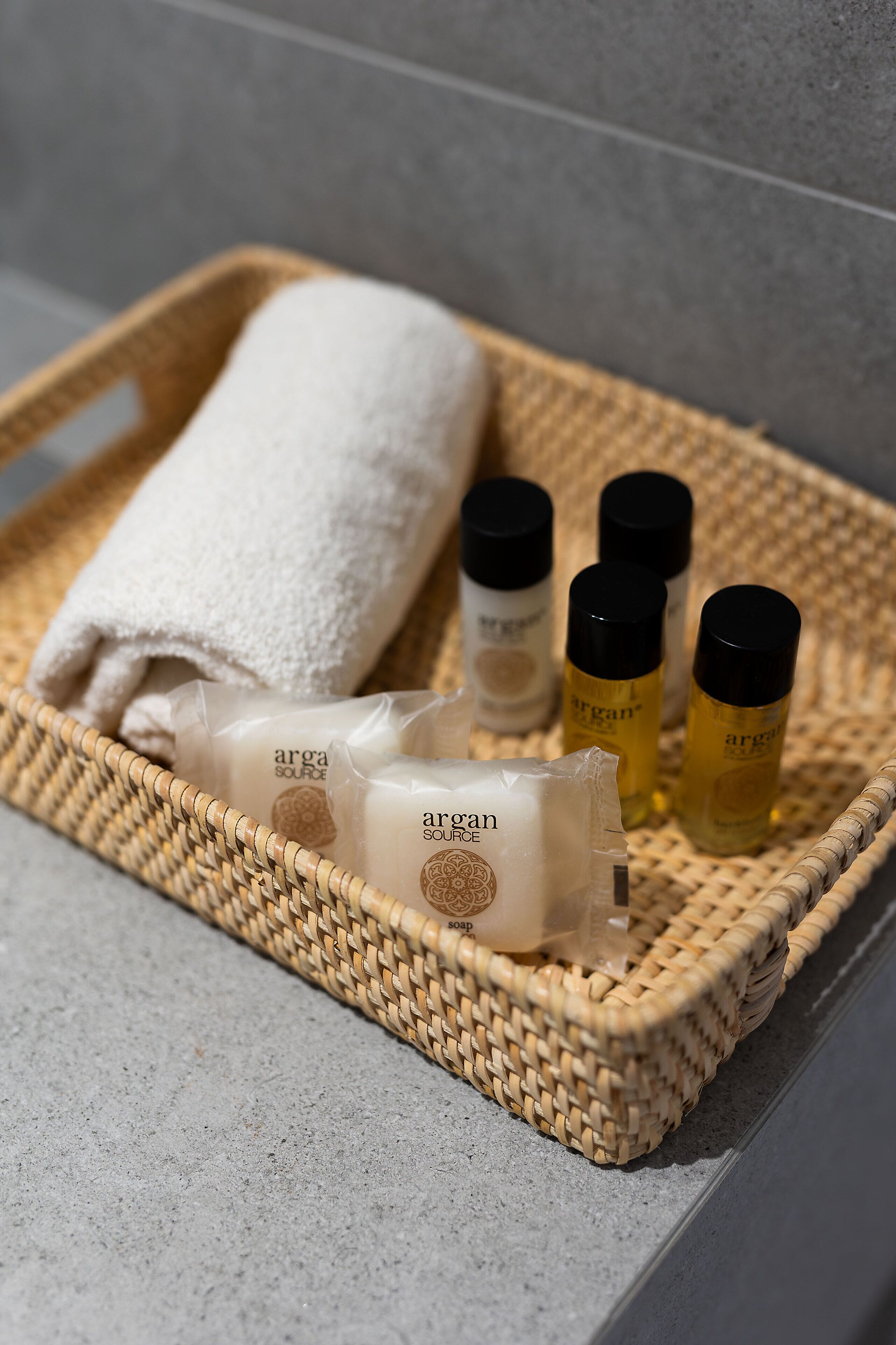 A wicker tray with a white towel, soap bars, and bottles of lotion on a tiled surface.