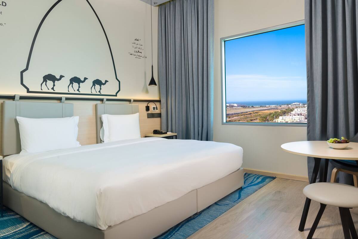 A hotel room with a large bed, a small round table with a bowl of fruit, a view of the city, and a wall decoration of camels.