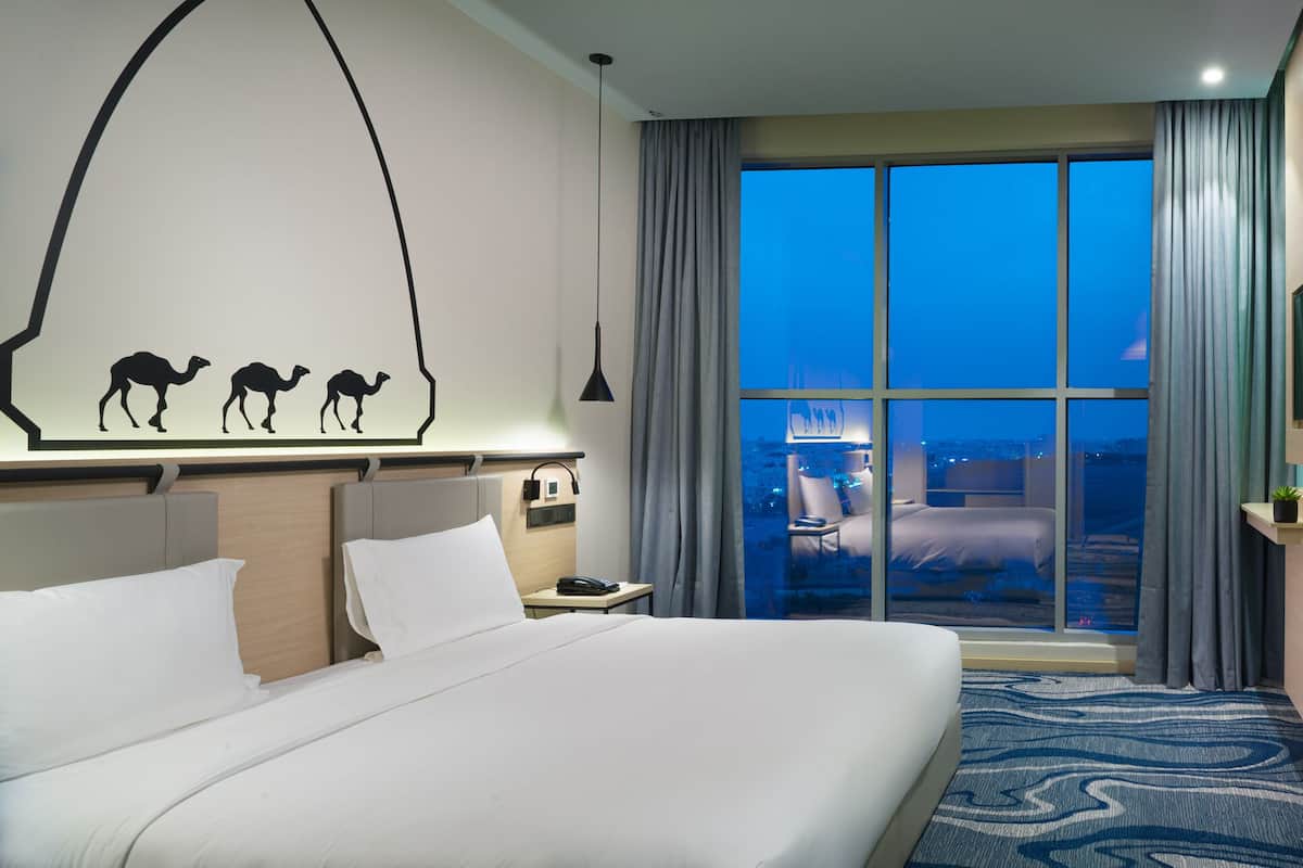 A hotel room with a large bed, a desk, a chair, a small table, and a view of the city at night.